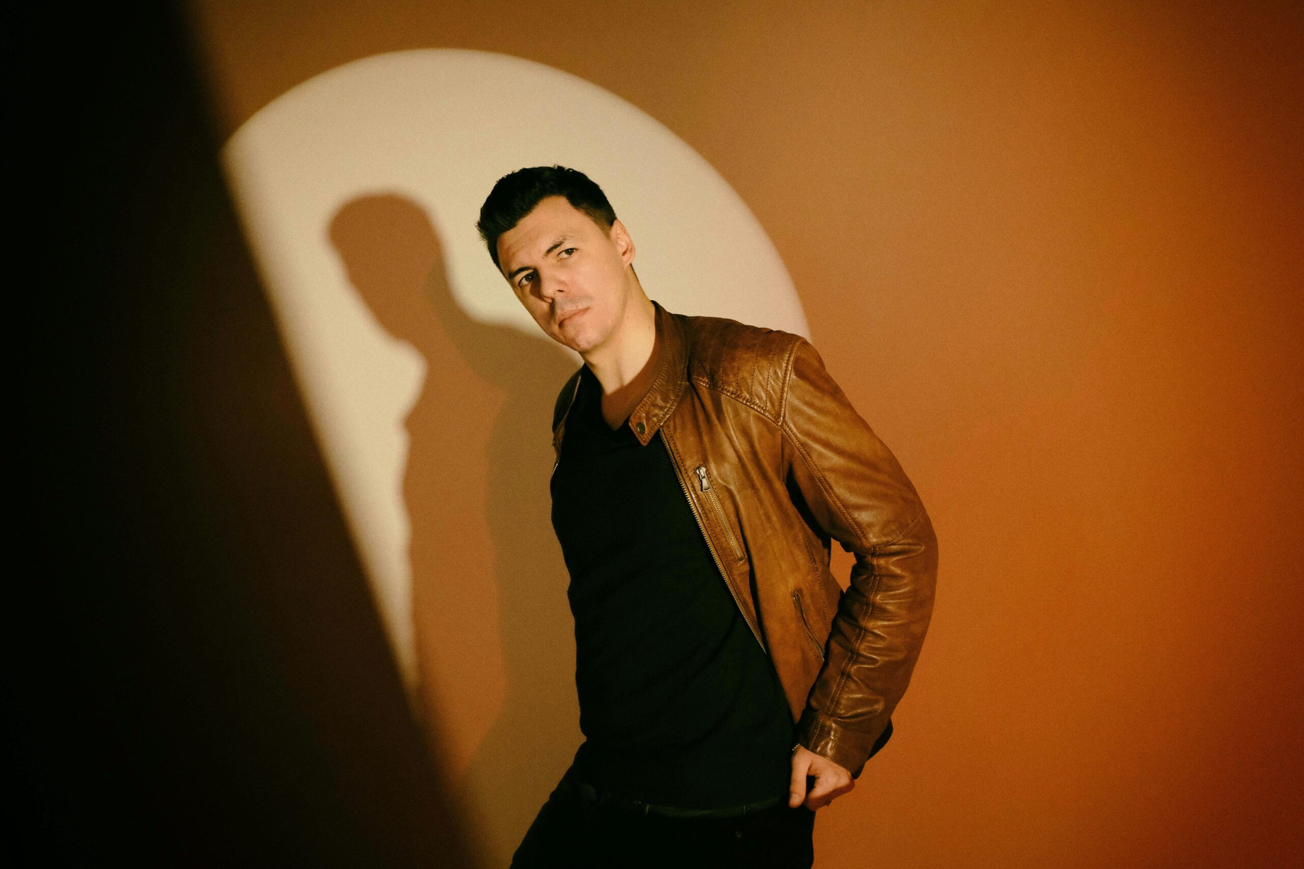 A fashionable man wearing a brown leather jacket posing confidently in a studio setting.